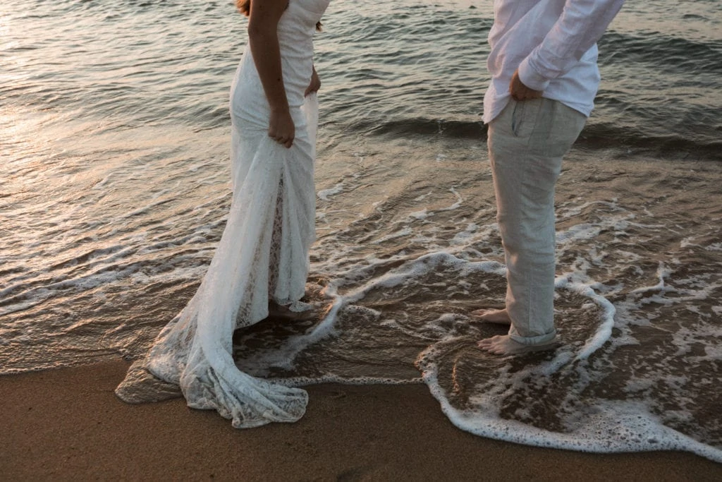 a bride and groom in their wedding attire stand at the edge of the beach getting their feet wet in the waves