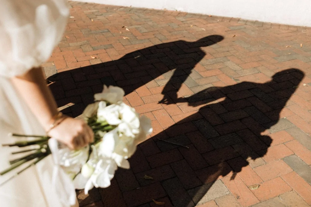in the foreground is the hand of a bride holding a wedding bouquet on her elopement in the background are the shadows of the bride and groom holding hands
