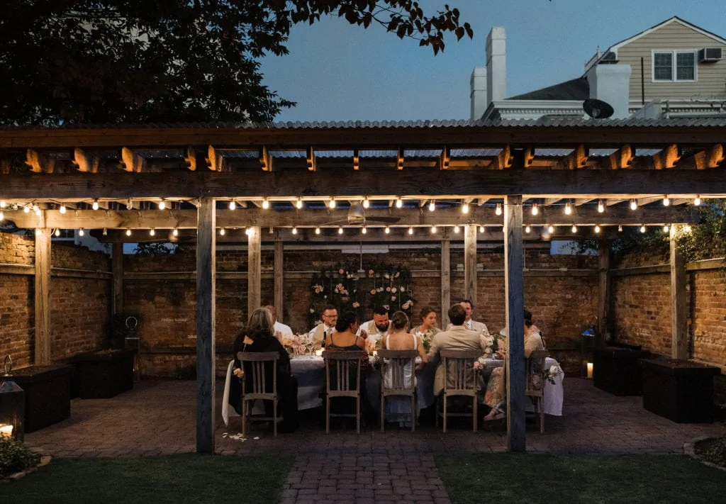 a bride and groom sit with their 9 guests on a patio underneath some twinkly lights at a table eating dinner at their elopement