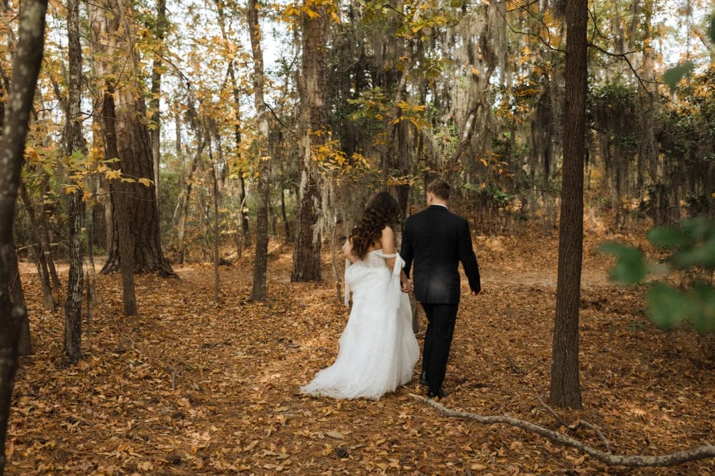 a bride and groom hold hands and walk through a spanish moss covered forest during fall