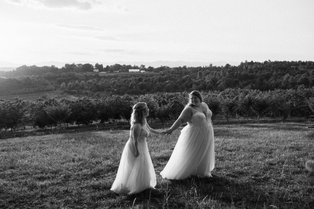 black and white photo of two brides wearing wedding gowns and walking, holding hands, and smiling at each other in a vineyard