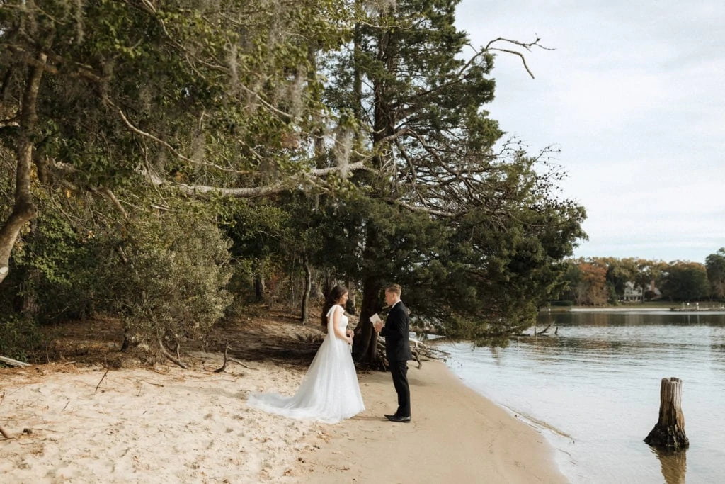 a couple in wedding attire stand at the edge of the water on the beach of first landing state park