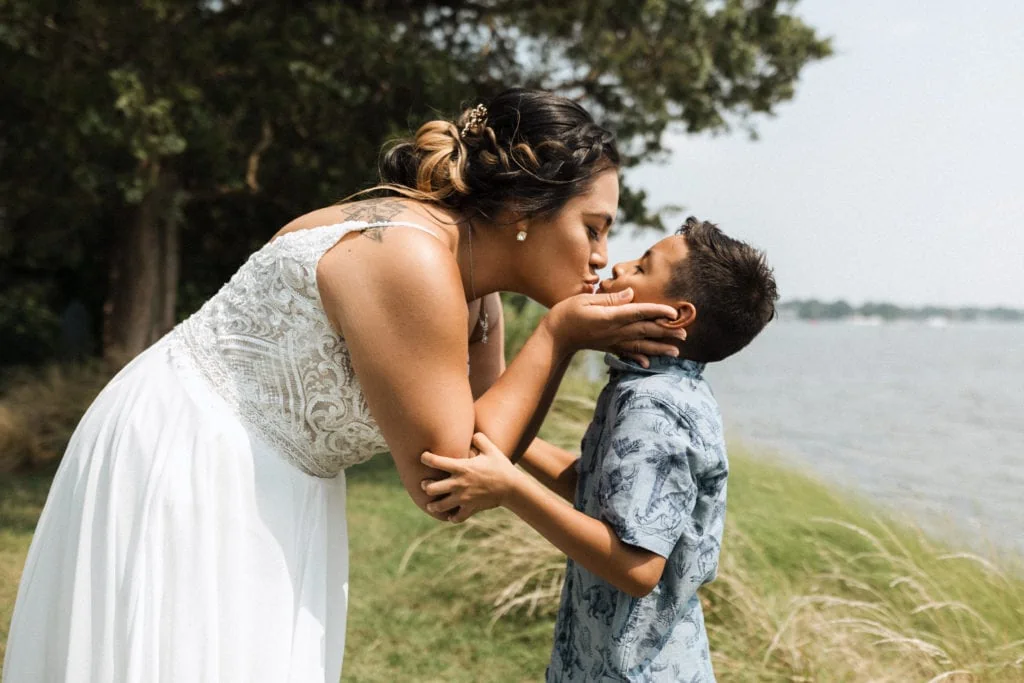 a mom in her wedding dress reaches down to give her son a kiss by the water