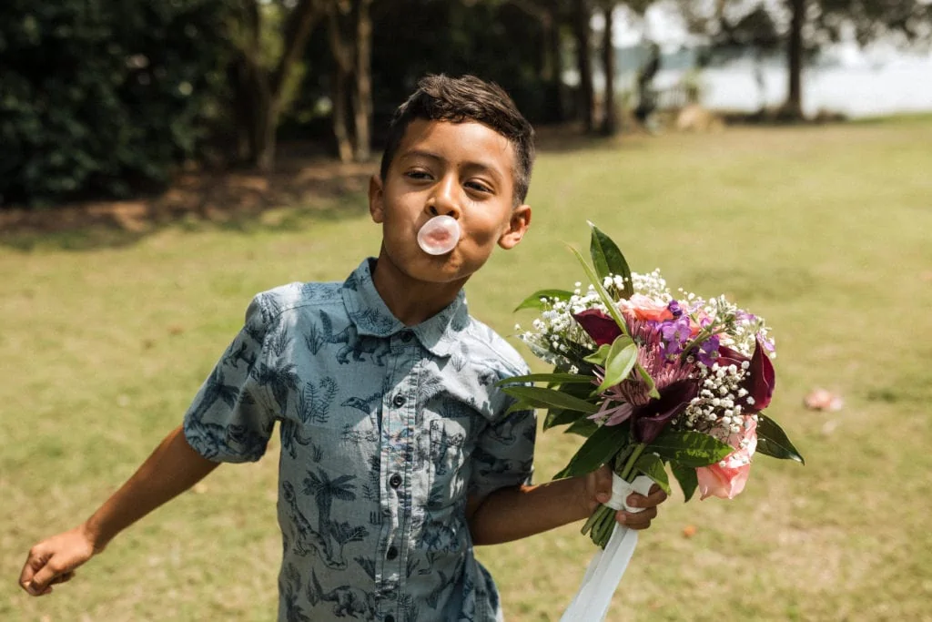 a young boy holds a wedding bouquet and blows a big pink gum bubble