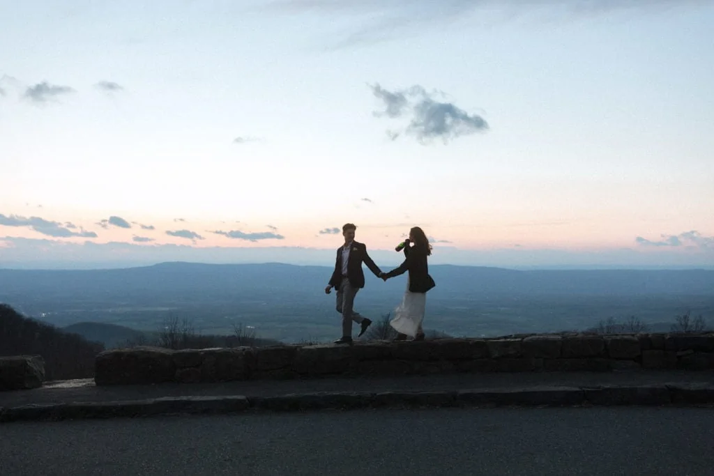 a bride and groom walk along Skyline Drive in the mountains at dusk drinking champagne