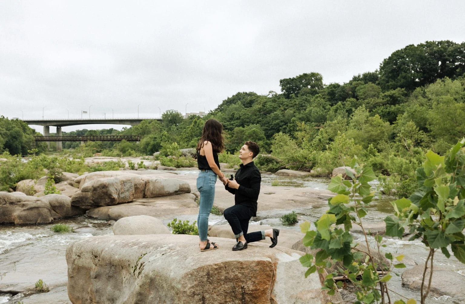 a man on one knee proposing to his girlfriend at Belle Isle in Richmond. He is on a big rock surrounded by river and trees.