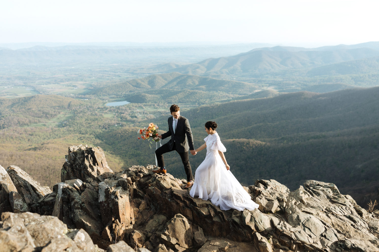 bride and groom walk along a mountain cliff ridge while holding bouquet