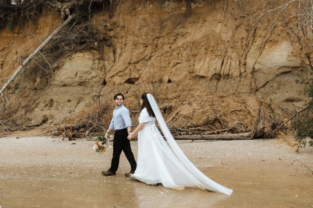 couple holding hands in wedding attire at the base of a cliff after having a wedding in a virginia state park by a cliff