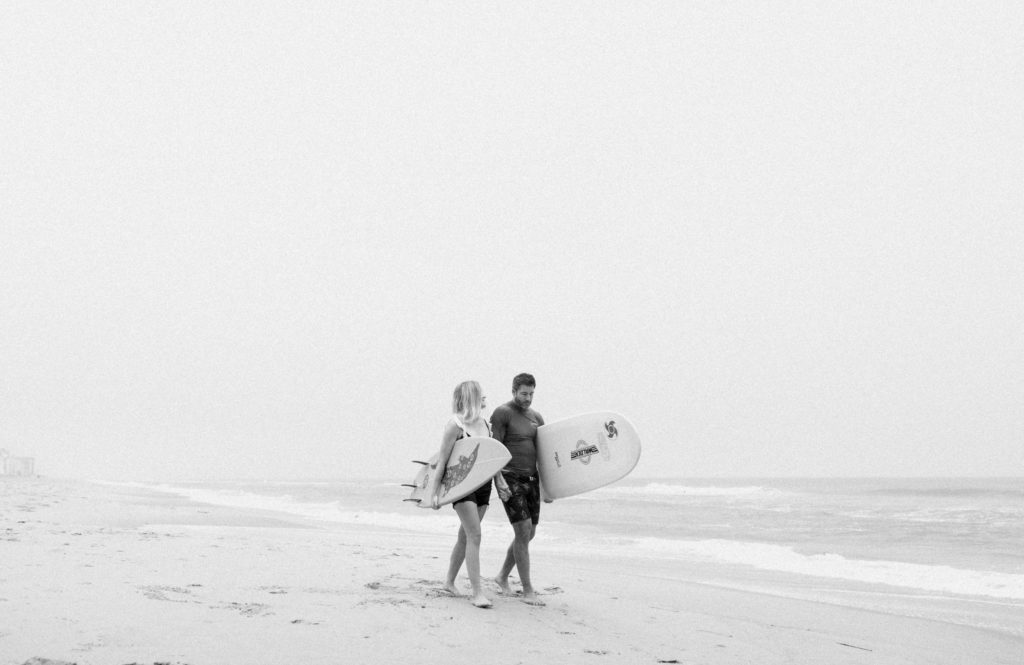 black and white photo of a couple holding hands at virginia beach and walking with their surfboards on a foggy day