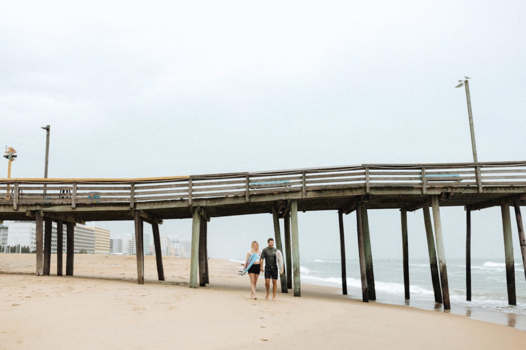 couple walking under the virginia beach fishing pier with their surfboards