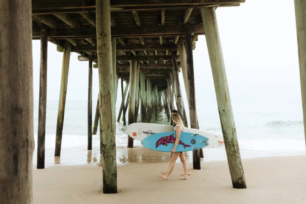 a couple with theirj surfboards walk under the virginia beach fishing pier