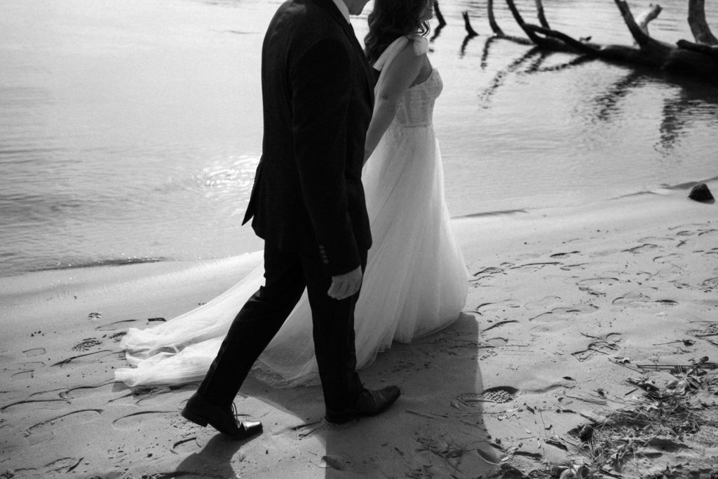 bride and groom holding hands walking through the sand at the water's edge on their elopement day