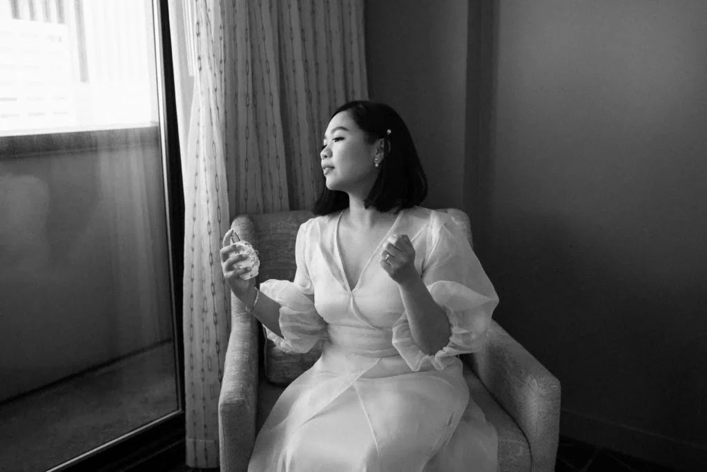 a bride sits by a window in her hotel room before the wedding putting on her perfume