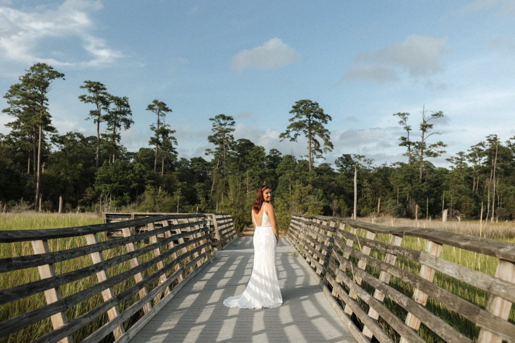 a bride stands on a boardwalk in her wedding gown surrounded by marsh and trees with blue sky