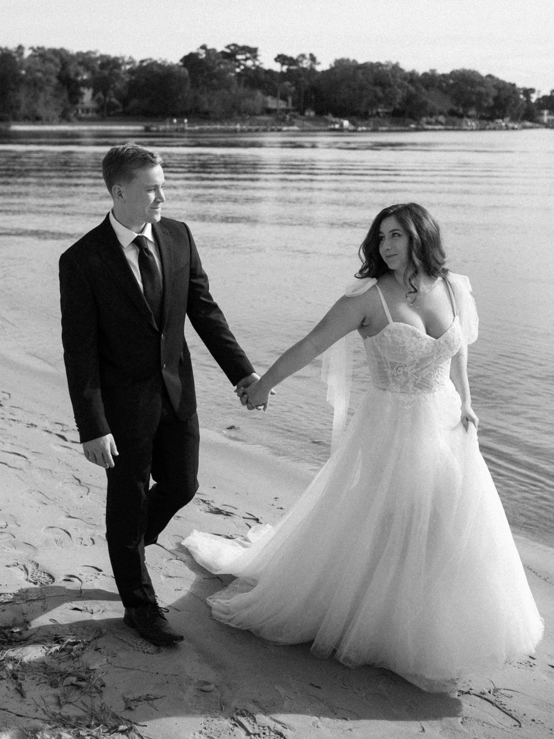 a bride and groom hold hands on the beach in their wedding attire