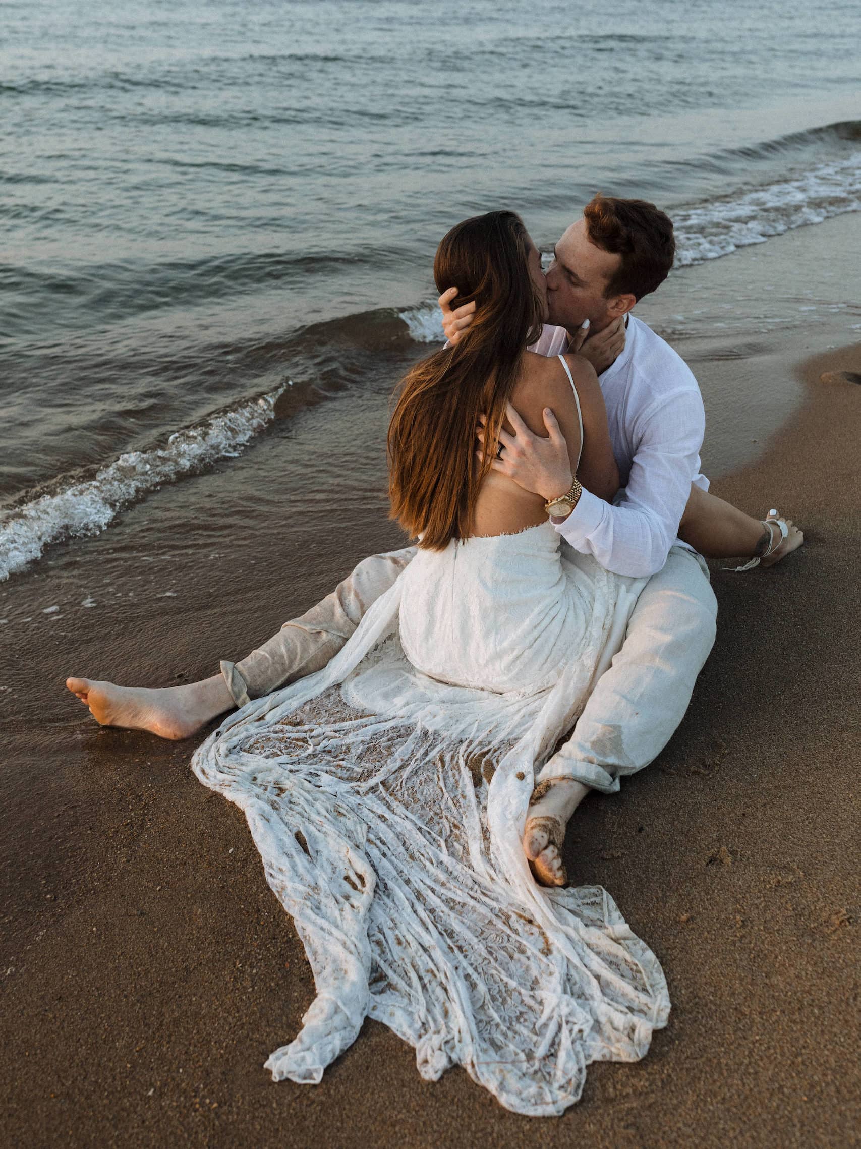 a couple in wedding attire sitting down and kissing on the sand as the waves come in