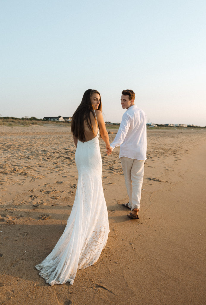 a bride and groom holding hands and walking along the beach