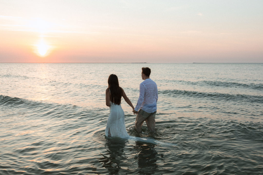 a bride and groom in the chesapeake bay holding hands in their wedding attire looking off into the sunset