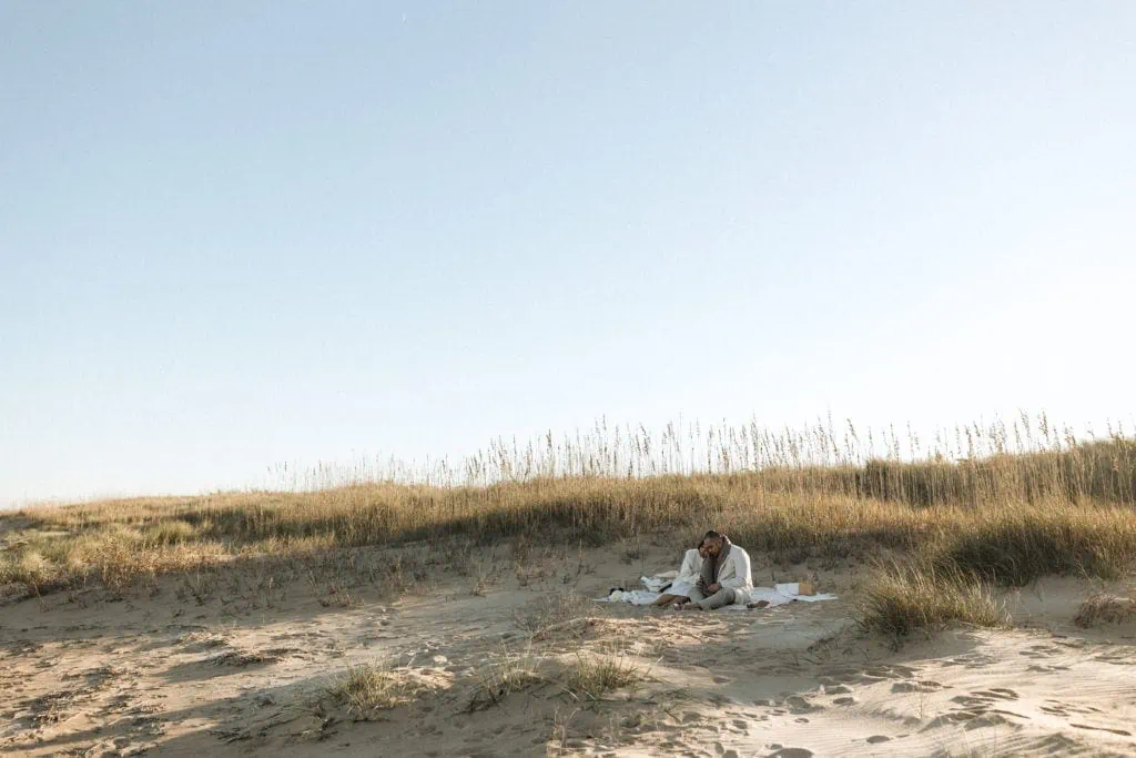 a bride and groom sit on a blanket near the sand dunes and hug