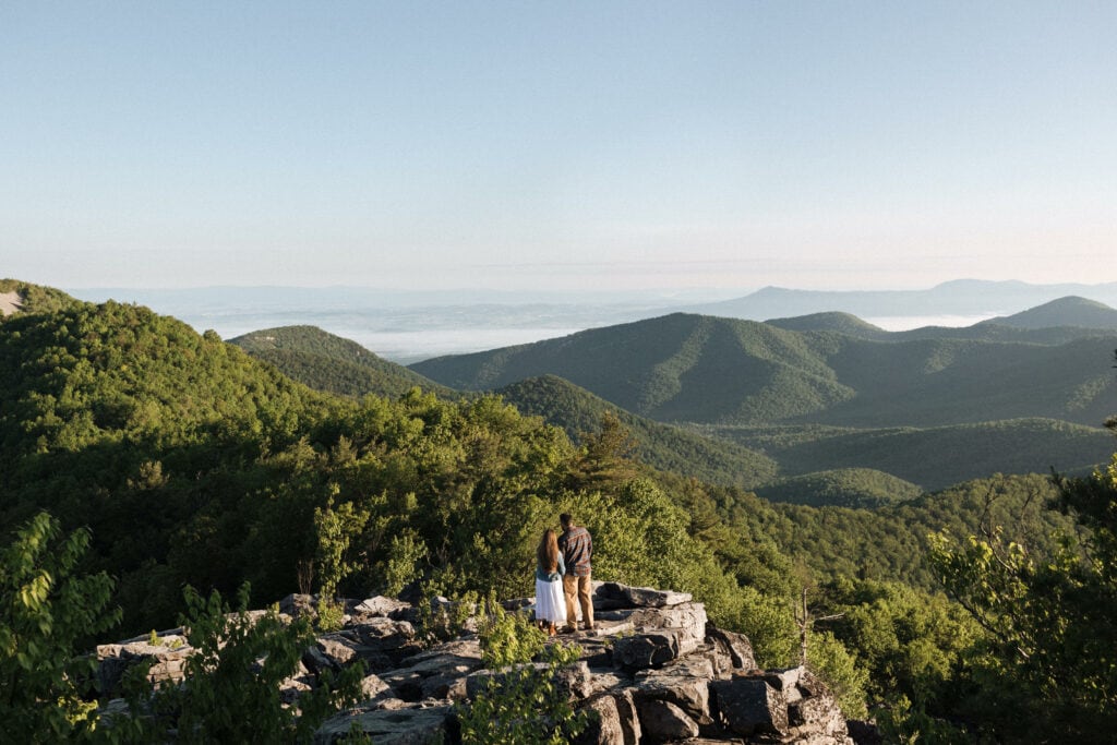a man and his wife stand on a rocky outcrop, hug and look at the blue ridge mountains 