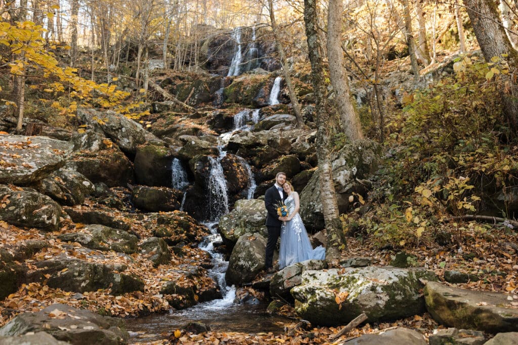 bride and groom smile and embrace in front of dark hollow falls, a waterfall, in shenandoah national park