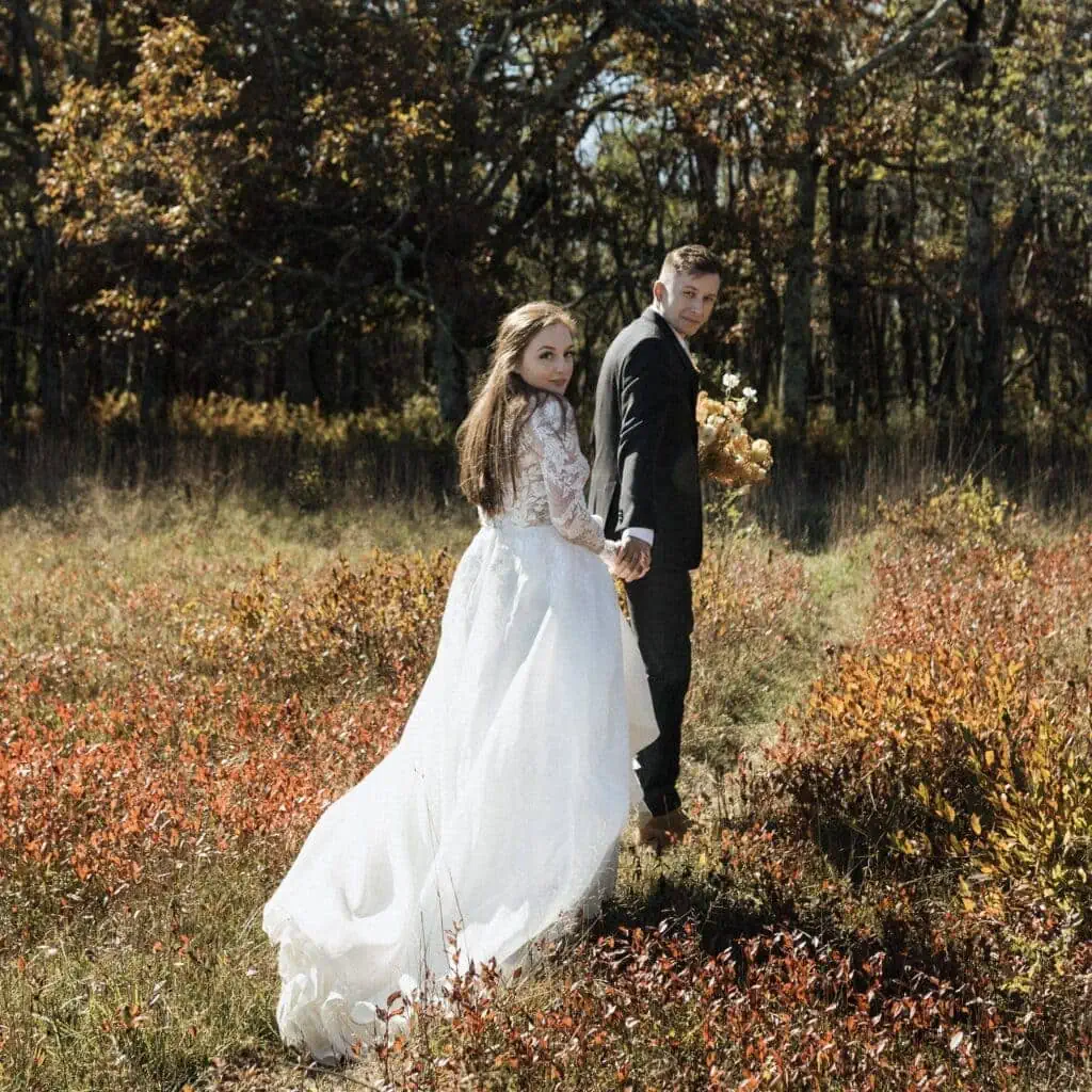 groom leads his bride through orange and red meadows in the fall
