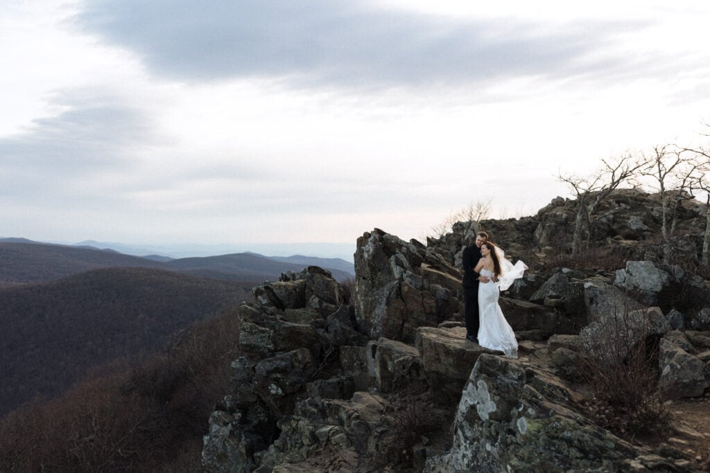 a bride and groom hug at sunrise on a cliff and look off in the distance at mountains
