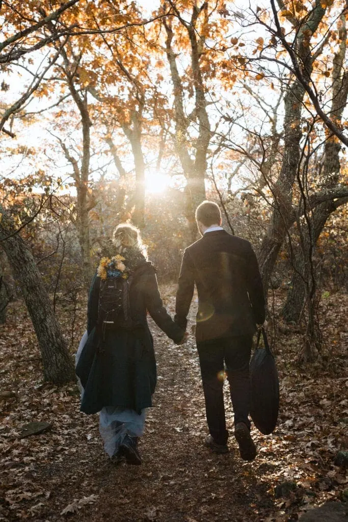 bride and groom holding hands walking through the forest with bouquet in backpack