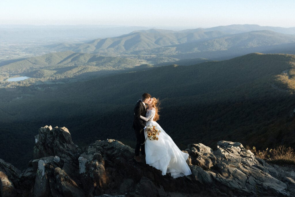 A bride and groom kissing at sunrise on the top of a mountain in Shenandoah National Park after their elopement wedding. The brides dress and hair blows in the wind and she holds a bright colored bouquet.