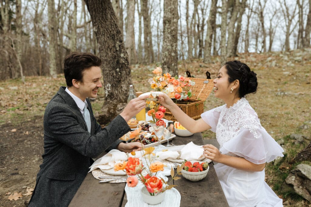 picnic in the woods a bride and groom feeding each other breakfast bagels surrounded by flowers