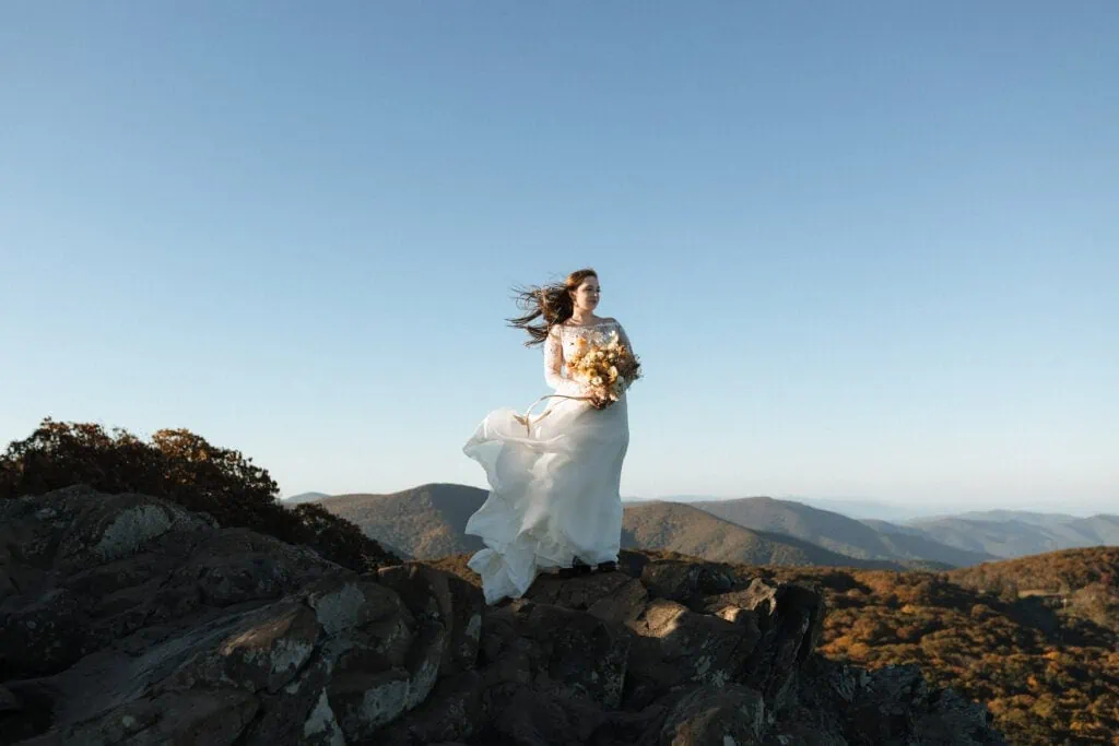 A bride stands on a mountain holding a wedding bouquet with her hair and dress blowing in the wind