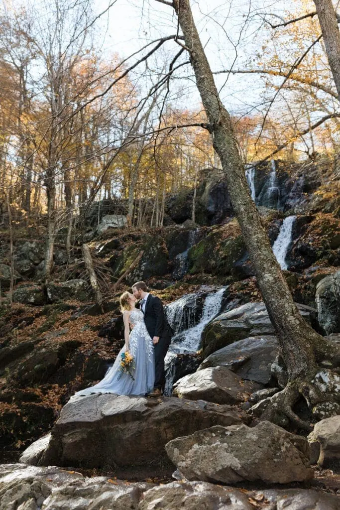 bride in blue dress kissing her groom in front of a waterfall surrounded by trees with yellow and orange leaves
