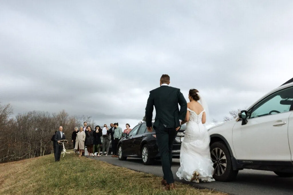 bride and groom walk past cars to meet their guests at a Skyline Drive overlook before their wedding ceremony in  a field with mountain views