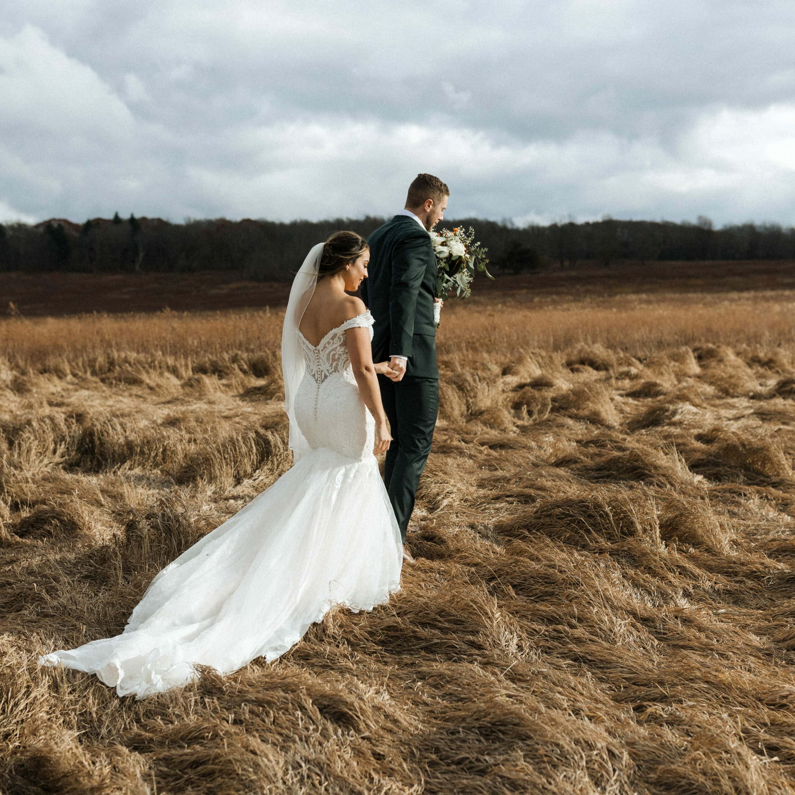 groom leads his bride through brown grasses in a beautiful open field the sky is stormy