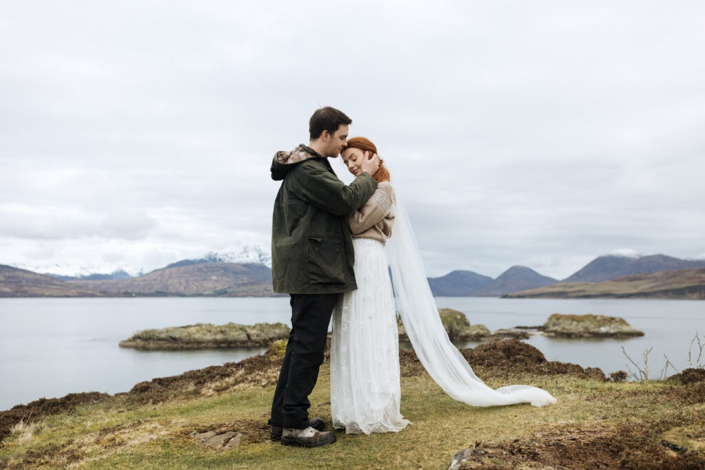 a bride and groom embrace on a cliff on the Isle of Skye with mountains and a lake in the background