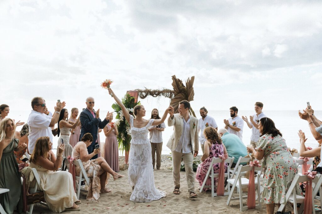 bride and groom smiling and raising arms up after just getting married on the beach and walking back down the aisle with their family and friends cheering on either side
