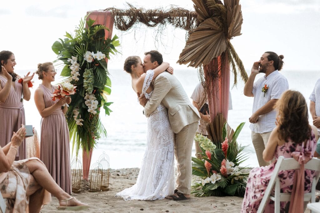 a bride and groom share their first kiss on the beach with a tropical floral arch in the background while their guests cheer them on